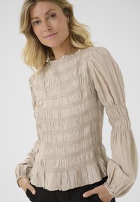 Blusa beige con un design texturizzato e arricciato, maniche lunghe a sbuffo e orlo a volant. Realizzata in tessuto leggero.