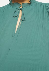 Błyszcząca niebieska bluzka z plisowaniem, z falbanami przy kołnierzyku, dekoltem w kształcie łezki oraz wiązaną wstążką. Lekki, zwiewny materiał o pionowej teksturze.