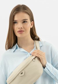 Borsa a tracolla in pelle beige con finitura testurizzata. Presenta una chiusura con zip e un sottile accento con logo, indossata sopra una camicia a righe blu chiaro.