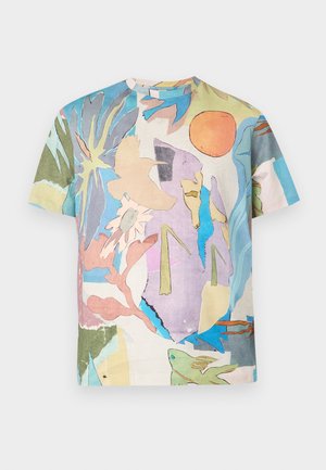 Muž s krátkymi blond vlasmi má na sebe farebné tričko s abstraktným vzorom, tmavohnedé nohavice a hnedé spoločenské topánky, stojí pred jednofarebným pozadím.