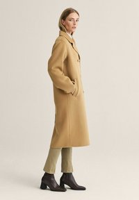 Beige uldfrakke med skræddersyet design, en-knaps lukning, klaplommer og en afslappet pasform. Sammen med olivengrønne bukser og brune ankelstøvler.