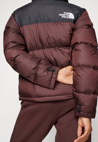 Doudoune bordeaux avec épaules noires, arborant un logo, un design matelassé, des poignets élastiques et un ourlet arrondi. Tissu texturé avec une finition lisse.