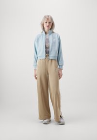 Veste zippée bleu clair avec des poignets côtelés, associée à un pantalon beige ample avec des boutons-pression sur le côté. Des baskets grises complètent la tenue.