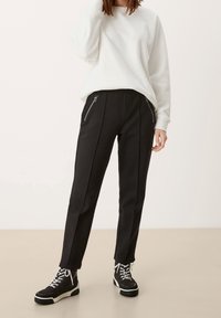 Pantaloni neri a sigaretta con cuciture laterali a contrasto e tasche anteriori con zip, abbinati a una felpa bianca con collo rotondo e sneakers nere.
