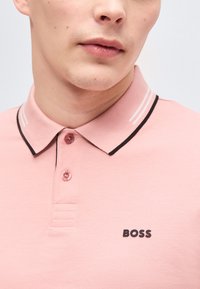 Άνδρας που φοράει ροζ πόλο μπλουζάκι με γιακά σε μαύρο περίγραμμα και δύο κουμπιά, με το λογότυπο "BOSS" στο στήθος.
