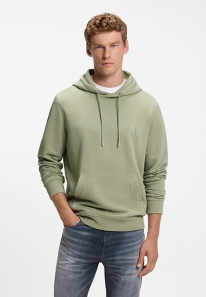 Homme aux cheveux bouclés portant un sweat à capuche vert clair et un jean bleu délavé, debout avec une main dans la poche sur fond blanc.
