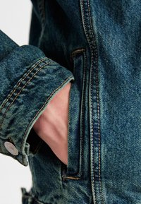 Detail van een spijkerjasje met een hand in een zak. De stof is vervaald blauw met zichtbare stiksels en een metalen knoop als accent.