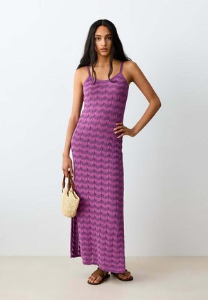 Femme aux longs cheveux foncés portant une robe longue violette à motifs et des sandales marron, tenant un petit sac à main tissé.