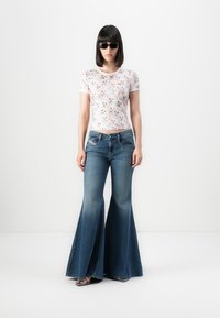 Kvinde iført sorte solbriller, hvid blomstret kortærmet top, brede blå jeans og sorte åbne hæle, stående mod en hvid baggrund.