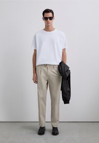 Man wearing black sunglasses, white t-shirt, beige pants, black shoes, holding black leather jacket in right hand against plain wall.