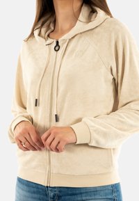 Felpa con zip color beige, dalla texture morbida, con cappuccio regolabile e dettagli metallici neri. Presenta maniche raglan e polsini a coste all'orlo.