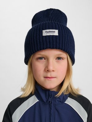 Kind met blond haar dat een donkerblauwe gebreide muts met het label "Hummel Sportswear" draagt en een donkerblauwe sportjas met grijze en zwarte accenten.