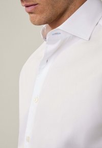 Witte overhemden met een klassieke kraag, voorzien van een kleine knopenlijst en lichtblauwe stiksels nabij de kraag. Effen textuur.