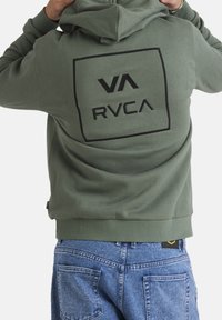 Dark green hoodie with a black rectangular logo featuring 'VA' and 'RVCA' on the back, paired with light blue denim jeans.