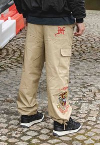 Beiga cargobyxor med avslappnad passform, med röda broderade mönster och sidofickor, i kombination med svarta sneakers och en bakgrund av texturerade stenar.