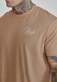 Light brown cotton t-shirt with a crew neckline, short sleeves, and a white logo on the left chest. Smooth texture, fitted design.