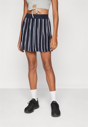 Vrouw draagt een marineblauwe rok met witte verticale strepen, een grijs cropped top, witte sokken en zwarte sneakers, staand tegen een effen achtergrond.