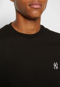 Black cotton t-shirt with a round neckline, featuring a small white embroidered logo on the left chest area. Smooth texture.