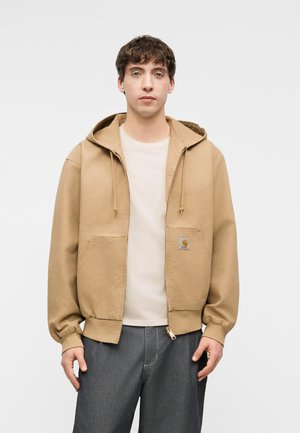 Ung mand iført en beige hættetrøje med lynlås over en hvid T-shirt og grå bukser, stående mod en enkel hvid baggrund.