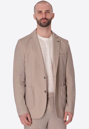 Uomo con barba corta che indossa giacca beige, pantaloni e camicia bianca, in piedi su uno sfondo chiaro e uniforme.