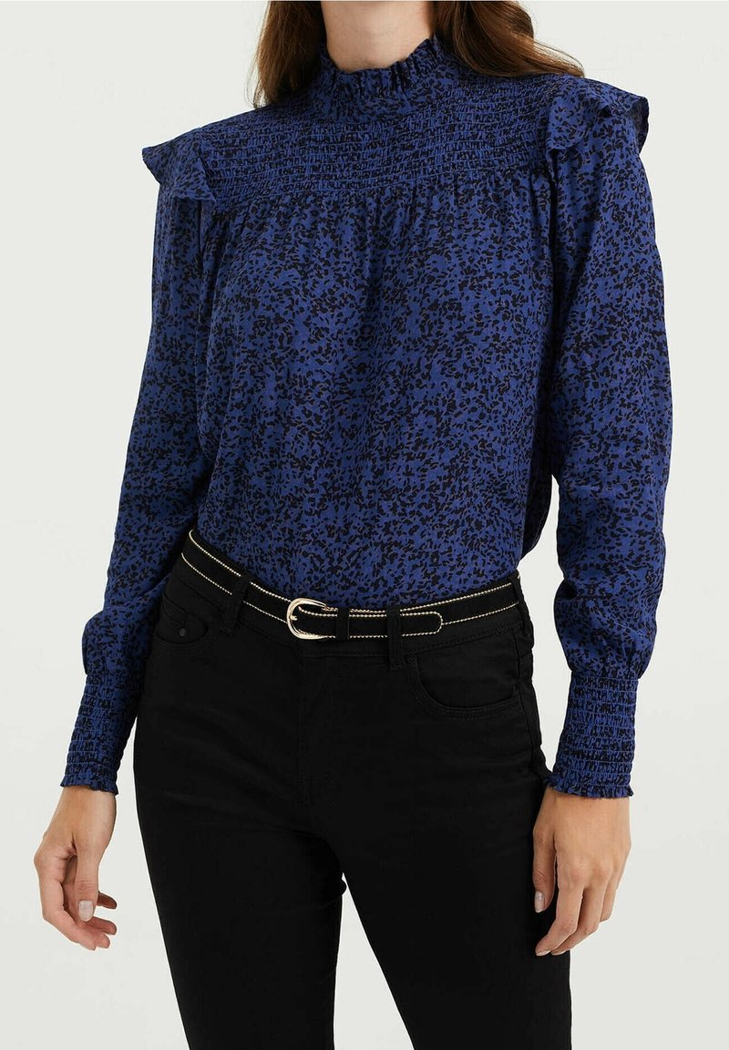 Žena v modré vzorované halence s vysokým límečkem, smockovanými rameny a manžetami, zastrčené do černých kalhot s černým páskem.