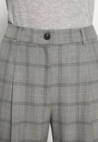 Szare spodnie w kratę z czarnym zapięciem na guzik, charakteryzujące się dopasowanym pasem, fałdami oraz cienkimi, jasnymi liniami wplecionymi w wzór.