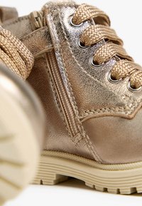 Metallisch goldene Knöchelstiefel mit strukturiertem Finish, runden Schnürsenkeln, seitlichem Reißverschluss und einer beige Gummisohle mit geriffeltem Profil.