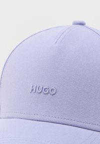 Απαλό μωβ καπέλο μπέιζμπολ με κεντημένο λογότυπο «HUGO» στο κέντρο του μπροστινού πάνελ, που δείχνει την υφή του υφάσματος και τη λεπτομέρεια του επάνω κουμπιού.