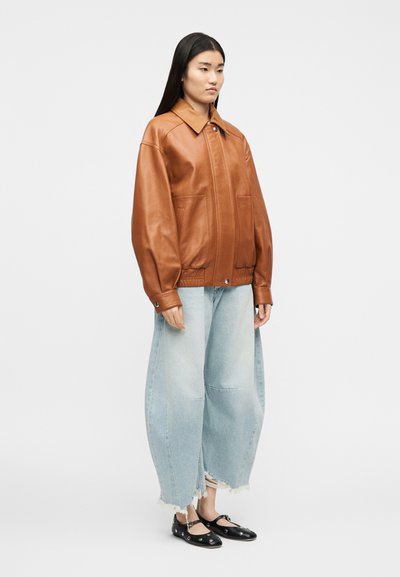 Γυναίκα που φοράει καφέ δερμάτινο μπουφάν, φαρδιά ανοιχτό μπλε τσαλακωμένα τζιν και μαύρα παπούτσια Mary Jane με ασημί τρύπες, στέκεται μπροστά σε λευκό φόντο.