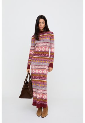 Frau trägt ein langes, mehrfarbig gemustertes Kleid mit langen Ärmeln, hält eine große dunkelbraune Handtasche und trägt beige Stiefeletten.