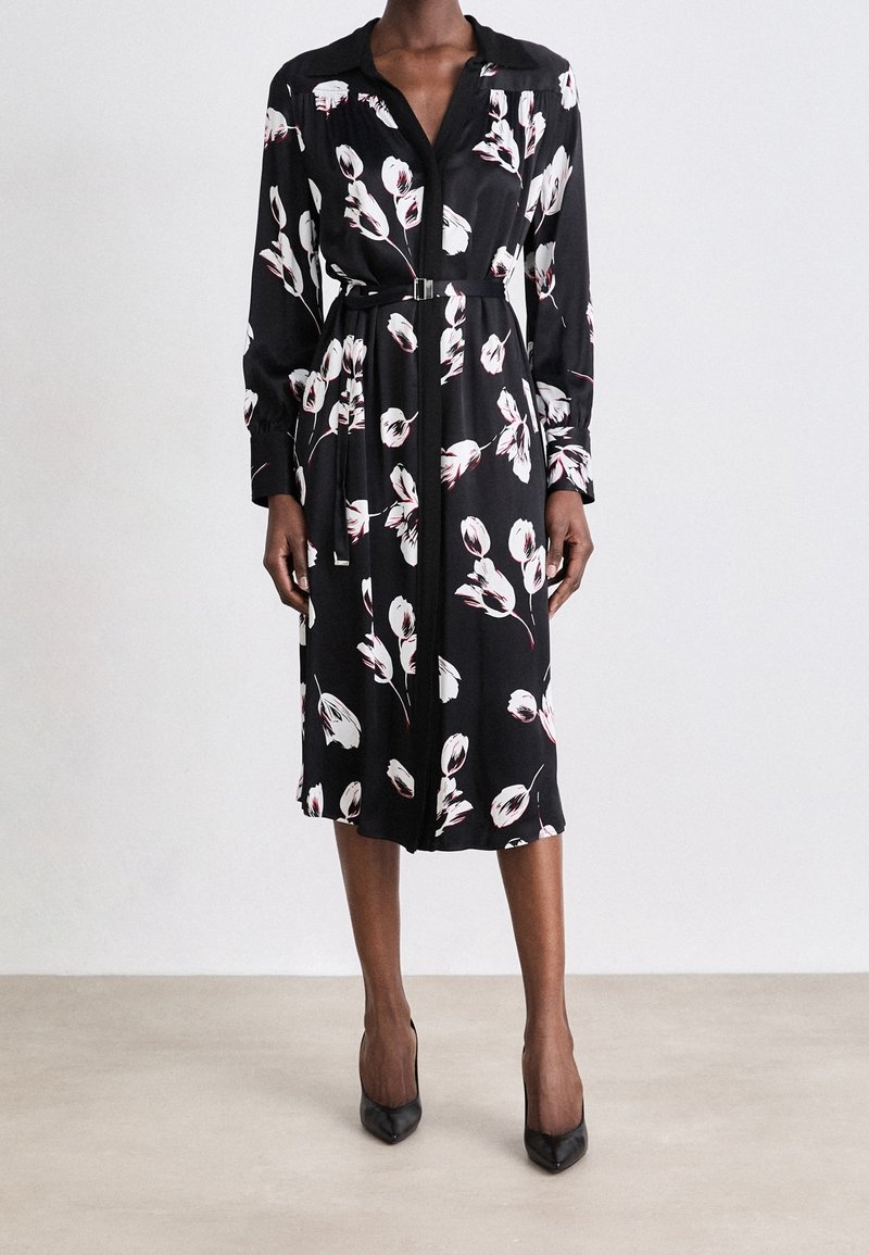 Femme portant une robe midi noire avec imprimé floral blanc, manches longues, col, boutons sur le devant, ceinture à la taille et talons noirs à bout pointu.