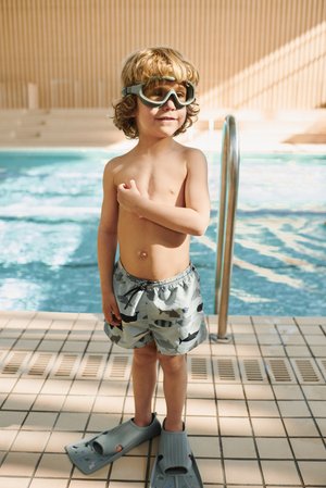Αγόρι που φοράει ανοιχτό μπλε μαγιό με σχέδια ψαριών, γκρι πτερύγια και γυαλιά κολύμβησης, στέκεται δίπλα σε μια πισίνα με πλακάκια στο πάτωμα.