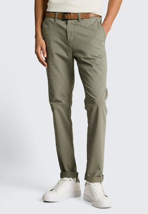 Homme portant un pantalon olive retroussé avec une ceinture marron et des baskets blanches, debout devant un fond clair uni.