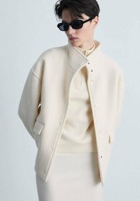 Crème-kleurige wollen jas met een rechtopstaande kraag, drukknoopsluitingen en zijzakken, gelaagd over een lichtbeige geribd topje en rok.