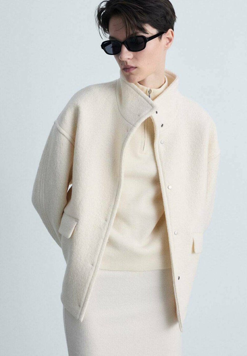 Crème-kleurige wollen jas met een rechtopstaande kraag, drukknoopsluitingen en zijzakken, gelaagd over een lichtbeige geribd topje en rok.
