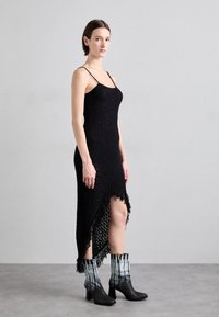 Vestido negro de punto texturizado con tirantes de espagueti y dobladillo asimétrico. Combinado con botas negras hasta el tobillo que presentan acentos de patrón plateado.
