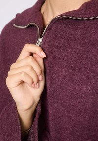 Bordeauxfarbener Fleece-Reißverschluss mit strukturiertem Finish, ausgestattet mit einem silbernen Reißverschluss am Kragen und einer Hand, die den Reißverschlussgriff hält.