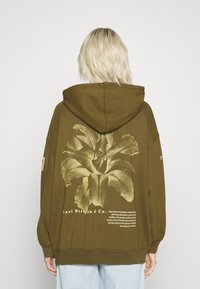 Persona con cabello rubio que lleva una sudadera color verde olivo con un gran gráfico de lirio amarillo y el texto de Levi Strauss & Co. en la parte posterior.