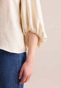 Top color crema con maniche a sbuffo, elastico ai polsini e una consistenza morbida. Abbinato a un denim blu scuro, che mostra una silhouette fluida.