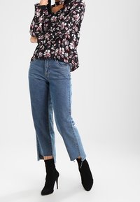 Blouse noire à motifs floraux avec manches longues, associée à un jean court bleu clair aux ourlets effilochés et des bottines noires à talons hauts.