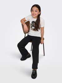 Camiseta blanca con diseño gráfico negro, pantalones negros de pierna recta y mocasines negros brillantes, sentado en una silla de madera contra un fondo claro.