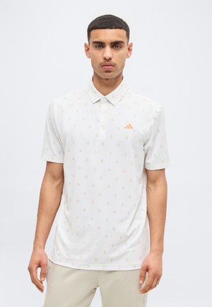 Fiatal férfi fehér, mintás galléros pólóban, narancssárga logóval, egyszínű világos háttér előtt áll.