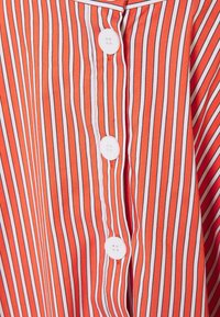 Oranssi raidallinen paita, jossa on pystysuoria viivoja eri leveyksissä, valkoisella kauluksella ja valkoisilla napilla edessä, valmistettu kevyestä kankaasta.