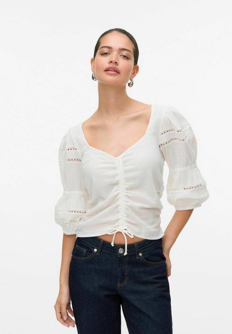 Blusa bianca con maniche a palloncino e scollatura a V; presenta un cordoncino in vita e dettagli in pizzo sulle maniche; tessuto morbido e strutturato.