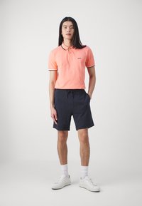 Šviesiai oranžinis polo marškinėliai su juodu apvadu, dėvimi su tamsiai mėlynomis šortais. Modelis vilki baltus sportinius batelius ir sportines kojines, stovėdamas prieš paprastą foną.