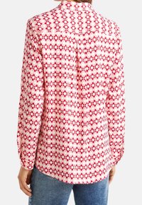 Chemise à manches longues rouge et blanche avec un motif géométrique. Le tissu semble lisse ; elle possède un col et des poignets à boutons, avec un ourlet incurvé.