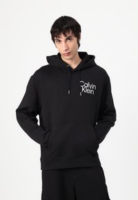 Black hoodie with a front kangaroo pocket, drawstring hood, and white "Calvin Klein" logo on the left side, made of soft fabric.
