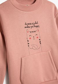 Sudadera rosa con cuello acanalado, que presenta un bolsillo frontal y un gráfico de un gato sonriente con el texto "hace más de lo que te hace feliz."