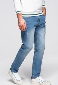 Jean en denim bleu clair à coupe slim, avec des détails usés et cinq poches, associé à des baskets blanches et vertes.