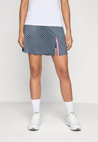 Femme portant une jupe de sport à motifs bleu marine avec une fente sur le côté laissant voir des shorts intérieurs, des chaussettes blanches, des baskets blanches et un haut blanc.